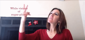 Figure 3 shows me, a young woman with shoulder-length brown hair, wearing a red long-sleeve shirt with stockings on a chimney behind me. I am signing VISIONS and looking upward towards red captions that appear in the upper left corner. My eyes and signs are directed towards the space that the captions appear in. The captions read “While visions of sugar-plums.”