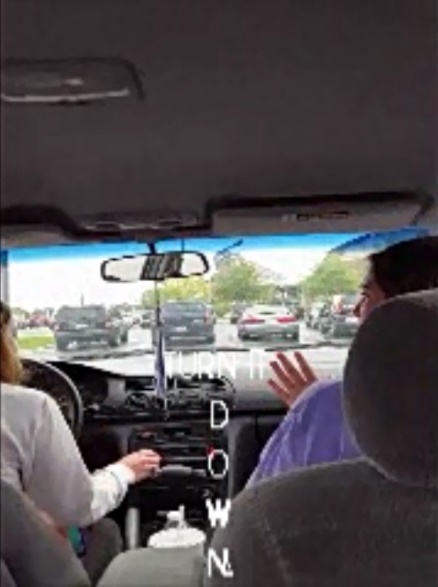 The camera is in the backseat of a car in the middle seat to show the front row of the car from the back. Two young women are seated in the front seats and looking at each other, so only their profiles are seen. The driver has her hand on the radio button as captions appear at the top of the screen. The captions read “TURN IT DOWN” in white capitalized letters and the word “DOWN” appears as a vertically written word. Each letter in the word DOWN appears alone so that the viewer’s eye will go down the screen to read the word.